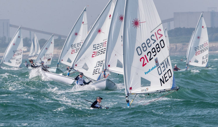 Flotte de dériveurs Laser en course, voiles blanches avec codes pays visibles, navigateurs contrebalançant, sur mer agitée avec ligne côtière en arrière-plan.